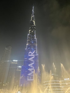 A towering skyscraper illuminated with bright lights, dominating the skyline at night. The building features the word 'EMAAR' displayed prominently in glowing letters. The scene includes a well-lit fountain in the foreground, with water jets creating an elegant pattern against the dark sky.