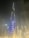 A towering skyscraper illuminated with bright lights, dominating the skyline at night. The building features the word 'EMAAR' displayed prominently in glowing letters. The scene includes a well-lit fountain in the foreground, with water jets creating an elegant pattern against the dark sky.