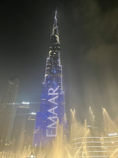 A towering skyscraper illuminated with bright lights, dominating the skyline at night. The building features the word 'EMAAR' displayed prominently in glowing letters. The scene includes a well-lit fountain in the foreground, with water jets creating an elegant pattern against the dark sky.