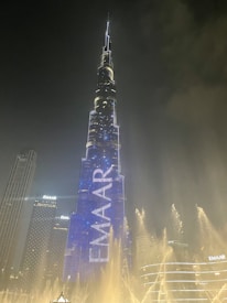 A towering skyscraper illuminated with bright lights, dominating the skyline at night. The building features the word 'EMAAR' displayed prominently in glowing letters. The scene includes a well-lit fountain in the foreground, with water jets creating an elegant pattern against the dark sky.