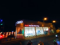 A modern car dealership with the brand Audi prominently displayed in neon lights. The building features large glass windows showcasing cars inside. The scene is captured at night, with streetlights illuminating the area and a few people and vehicles visible on the street.