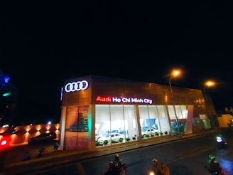 A modern car dealership with the brand Audi prominently displayed in neon lights. The building features large glass windows showcasing cars inside. The scene is captured at night, with streetlights illuminating the area and a few people and vehicles visible on the street.