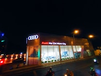 A modern car dealership with the brand Audi prominently displayed in neon lights. The building features large glass windows showcasing cars inside. The scene is captured at night, with streetlights illuminating the area and a few people and vehicles visible on the street.
