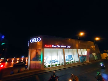 A modern car dealership with the brand Audi prominently displayed in neon lights. The building features large glass windows showcasing cars inside. The scene is captured at night, with streetlights illuminating the area and a few people and vehicles visible on the street.