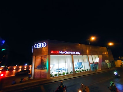 A modern car dealership with the brand Audi prominently displayed in neon lights. The building features large glass windows showcasing cars inside. The scene is captured at night, with streetlights illuminating the area and a few people and vehicles visible on the street.