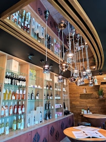 The image depicts a modern wine bar interior with wooden and metal decor. Shelves are filled with various bottles of wine, some in gift packaging. Overhead, there are hanging metallic pendant lights creating a sophisticated ambiance. The walls feature wooden paneling, and a small table with books or brochures is positioned in the foreground.