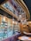 The image depicts a modern wine bar interior with wooden and metal decor. Shelves are filled with various bottles of wine, some in gift packaging. Overhead, there are hanging metallic pendant lights creating a sophisticated ambiance. The walls feature wooden paneling, and a small table with books or brochures is positioned in the foreground.
