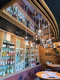 The image depicts a modern wine bar interior with wooden and metal decor. Shelves are filled with various bottles of wine, some in gift packaging. Overhead, there are hanging metallic pendant lights creating a sophisticated ambiance. The walls feature wooden paneling, and a small table with books or brochures is positioned in the foreground.