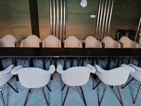 A conference room featuring two rows of houndstooth-patterned chairs neatly aligned around a long black table. The room has wooden paneling and metallic vertical panels on the walls. A wall clock is visible on the wall in the background.