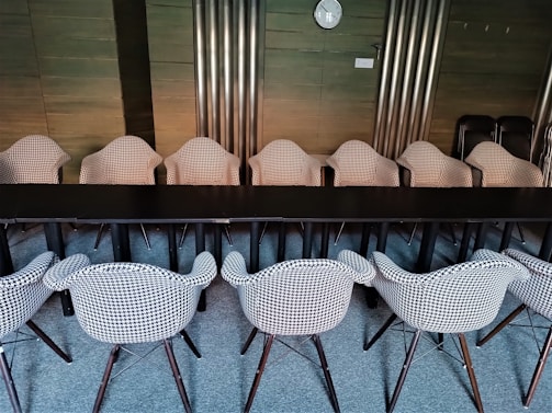 A conference room featuring two rows of houndstooth-patterned chairs neatly aligned around a long black table. The room has wooden paneling and metallic vertical panels on the walls. A wall clock is visible on the wall in the background.