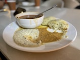 A white plate holds three steamed savory cakes alongside two types of chutneys, one creamy white with specks and another spiced brown. A small white bowl with sambar and a metal spoon sits on the edge of the plate.