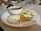 A white plate holds three steamed savory cakes alongside two types of chutneys, one creamy white with specks and another spiced brown. A small white bowl with sambar and a metal spoon sits on the edge of the plate.