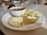 A white plate holds three steamed savory cakes alongside two types of chutneys, one creamy white with specks and another spiced brown. A small white bowl with sambar and a metal spoon sits on the edge of the plate.