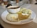 A white plate holds three steamed savory cakes alongside two types of chutneys, one creamy white with specks and another spiced brown. A small white bowl with sambar and a metal spoon sits on the edge of the plate.