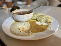 A white plate holds three steamed savory cakes alongside two types of chutneys, one creamy white with specks and another spiced brown. A small white bowl with sambar and a metal spoon sits on the edge of the plate.