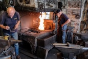 A blacksmith workshop featuring two men working. One man is focused on preparing materials near an anvil, while the other monitors a vibrant fire in a brick furnace. The room is filled with tools and machinery, giving it an industrial feel. Metal instruments hang from the walls, and the atmosphere is one of concentration and craftsmanship.