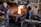 A blacksmith workshop featuring two men working. One man is focused on preparing materials near an anvil, while the other monitors a vibrant fire in a brick furnace. The room is filled with tools and machinery, giving it an industrial feel. Metal instruments hang from the walls, and the atmosphere is one of concentration and craftsmanship.