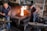 A blacksmith workshop featuring two men working. One man is focused on preparing materials near an anvil, while the other monitors a vibrant fire in a brick furnace. The room is filled with tools and machinery, giving it an industrial feel. Metal instruments hang from the walls, and the atmosphere is one of concentration and craftsmanship.