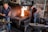 A blacksmith workshop featuring two men working. One man is focused on preparing materials near an anvil, while the other monitors a vibrant fire in a brick furnace. The room is filled with tools and machinery, giving it an industrial feel. Metal instruments hang from the walls, and the atmosphere is one of concentration and craftsmanship.
