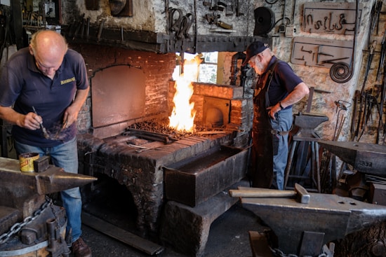 A blacksmith workshop featuring two men working. One man is focused on preparing materials near an anvil, while the other monitors a vibrant fire in a brick furnace. The room is filled with tools and machinery, giving it an industrial feel. Metal instruments hang from the walls, and the atmosphere is one of concentration and craftsmanship.