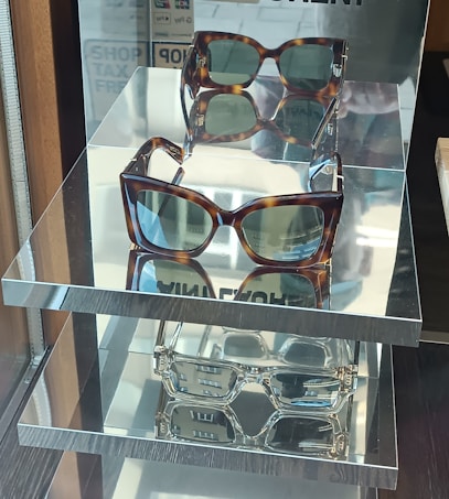 A display shelf with three pairs of sunglasses is positioned on mirrored surfaces, reflecting their image below. The sunglasses feature brown tortoiseshell frames with green-tinted lenses. The shelves are placed in a well-lit area, possibly near a window, contributing to reflections and a sleek presentation.