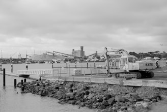 A construction vehicle is parked near a waterfront with an industrial area in the background. Piles of materials and cranes are visible across the water under a cloudy sky.