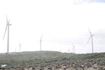 Several wind turbines are placed across a hilly landscape, surrounded by green vegetation and rocky terrain under a partly cloudy sky.