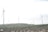 Several wind turbines are placed across a hilly landscape, surrounded by green vegetation and rocky terrain under a partly cloudy sky.