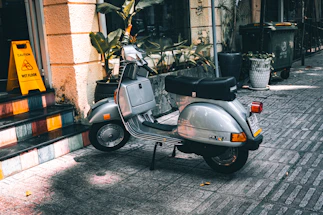 A silver scooter is parked on a tiled sidewalk next to a building with yellow walls. Nearby, a bright yellow caution sign indicates a wet floor. Potted plants and a large trash bin are visible in the background.