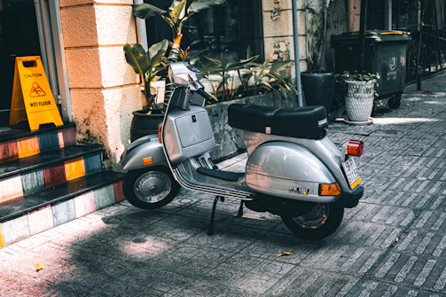 A silver scooter is parked on a tiled sidewalk next to a building with yellow walls. Nearby, a bright yellow caution sign indicates a wet floor. Potted plants and a large trash bin are visible in the background.