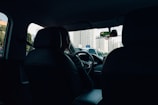 The inside of a car with a driver visible from the back seat, driving through a city with tall buildings visible through the windshield. The interior is dark, contrasting with the bright cityscape outside.
