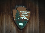 A wooden plaque features the National Park Service emblem, displaying a large sequoia tree, a mountain, and a bison against a rustic wooden background. The logo combines nature elements to signify conservation and wilderness areas.