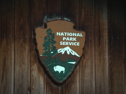 A wooden plaque features the National Park Service emblem, displaying a large sequoia tree, a mountain, and a bison against a rustic wooden background. The logo combines nature elements to signify conservation and wilderness areas.