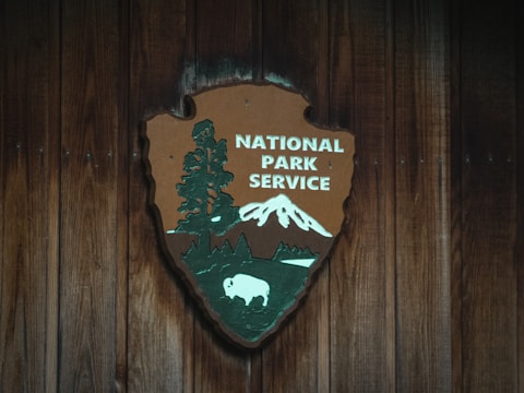 A wooden plaque features the National Park Service emblem, displaying a large sequoia tree, a mountain, and a bison against a rustic wooden background. The logo combines nature elements to signify conservation and wilderness areas.