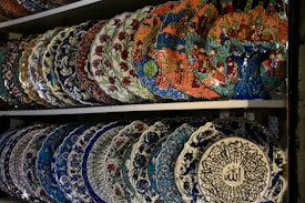 A collection of intricately designed ceramic plates with vibrant patterns and colors are displayed on a shelf. The plates feature floral motifs, traditional artistic designs, and detailed calligraphy. The top shelf contains plates with colorful, intricate scenes, while the bottom shelf features plates with bold blue patterns and calligraphy in the center.