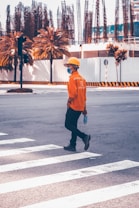 A person wearing an orange construction outfit, helmet, and face mask is crossing a road at a pedestrian crosswalk. They are holding a plastic water bottle in one hand. The background shows a construction site with palm trees and partially built structures surrounded by temporary fencing.