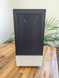 A sleek, modern computer case with a black perforated front panel and a white bottom section, placed on a wooden table. Green plant leaves appear in the background, adding a hint of nature to the scene.