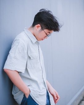 A person is standing with their back against a wall. They are wearing glasses and a light-colored shirt. Their head is slightly tilted downward, creating a thoughtful or introspective posture. The wall and background are a muted blue-gray color, adding to the subdued mood of the image.