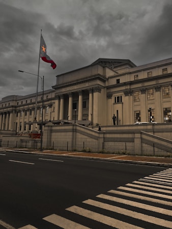 A grand neoclassical museum building with tall columns and an overcast sky. A national flag flies prominently in the foreground near a crosswalk and a wide street. The atmosphere is muted and historic, with a sense of architectural elegance.