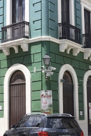 A corner of a green and white building with arched doors and black wrought-iron balconies. There is a vintage-style street lamp attached to the wall, surrounded by security cameras. A black Mercedes-Benz SLK 350 car is parked in front. A sign on the building indicates that the facility is open, mentioning 'Pirilo.'