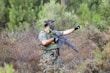 A hunter in camo gear aiming a rifle in a forest environment