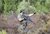 A person wearing camouflage gear, holding a rifle, and pointing towards something in the dense, green forest foliage. The individual is equipped with tactical gear and appears to be focused and alert.