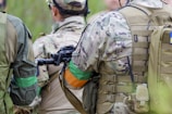 Several people wearing camouflage uniforms, tactical gear, and backpacks are standing closely together outdoors. They have colored arm bands, and one person is holding a rifle. The setting appears to be outdoor with lush greenery visible in the blurred background.