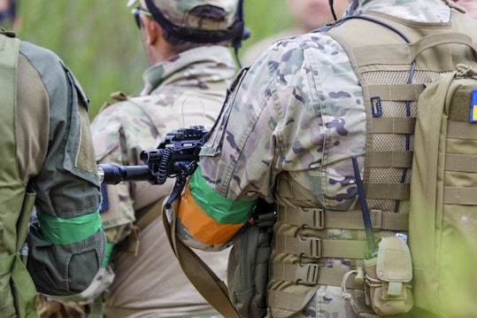 Several people wearing camouflage uniforms, tactical gear, and backpacks are standing closely together outdoors. They have colored arm bands, and one person is holding a rifle. The setting appears to be outdoor with lush greenery visible in the blurred background.