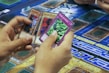 A person's hands holding four trading cards with colorful illustrations. The cards are being examined closely, and a game board featuring sections and text is visible in the background.