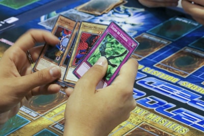 A person's hands holding four trading cards with colorful illustrations. The cards are being examined closely, and a game board featuring sections and text is visible in the background.