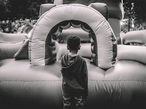 A child stands facing an inflatable play structure, which appears to be part of a bounce house setup. The scene is outdoors, with a crowd of people blurred in the background. The image is captured in black and white, highlighting textures and contrasts.