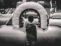 A child stands facing an inflatable play structure, which appears to be part of a bounce house setup. The scene is outdoors, with a crowd of people blurred in the background. The image is captured in black and white, highlighting textures and contrasts.