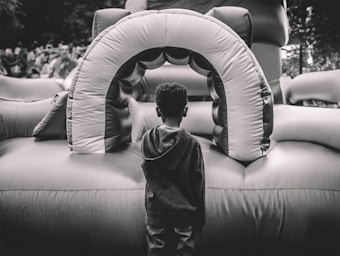 A child stands facing an inflatable play structure, which appears to be part of a bounce house setup. The scene is outdoors, with a crowd of people blurred in the background. The image is captured in black and white, highlighting textures and contrasts.