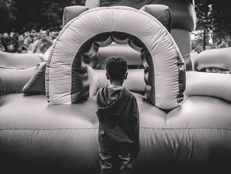 A child stands facing an inflatable play structure, which appears to be part of a bounce house setup. The scene is outdoors, with a crowd of people blurred in the background. The image is captured in black and white, highlighting textures and contrasts.